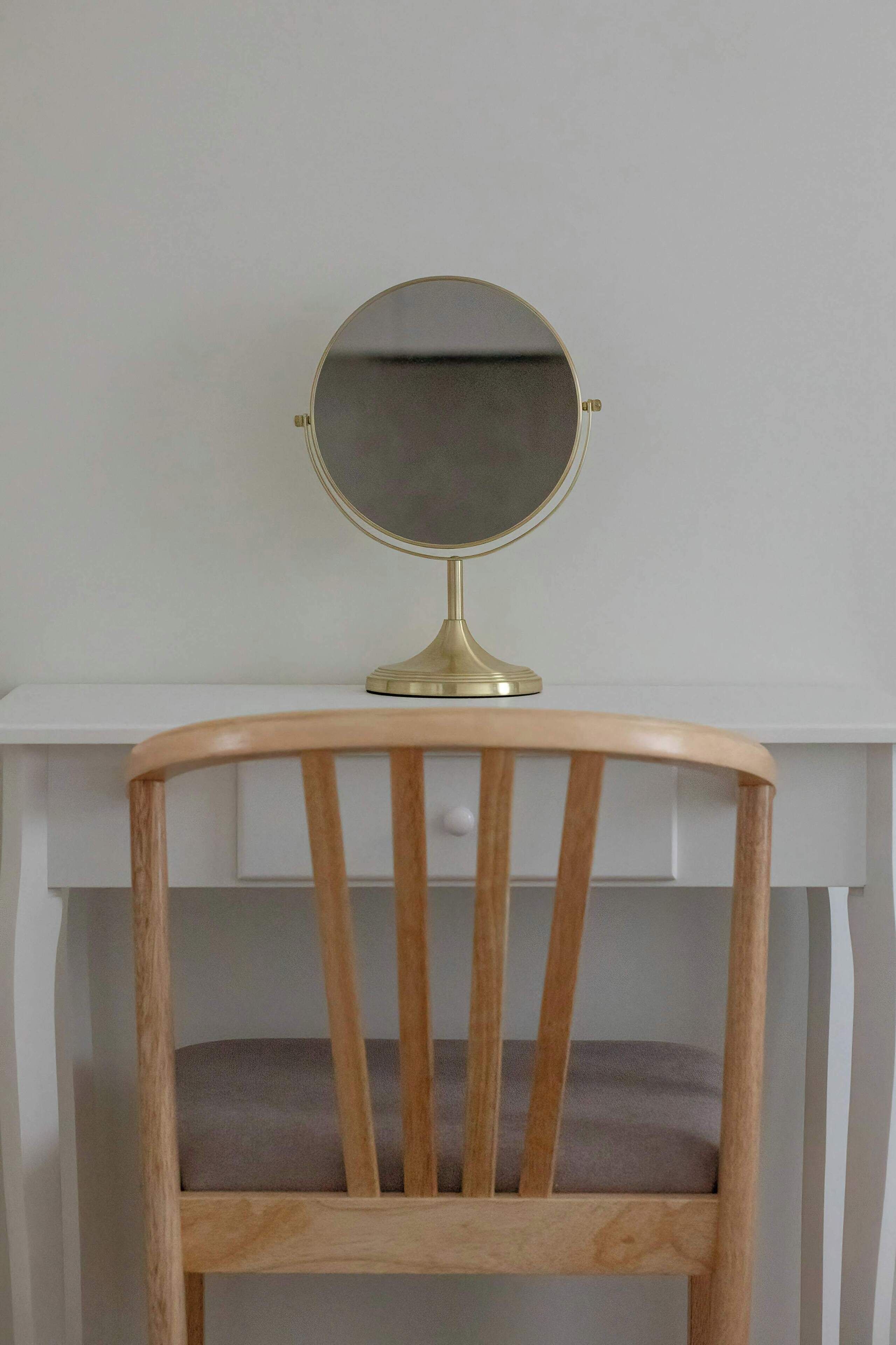 A wooden chair with a cushioned seat and backrest, positioned in front of a white desk with a round mirror on top.