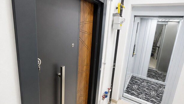 Un pasillo con una puerta oscura, un panel de madera, una pared blanca, una puerta blanca, una tubería blanca y un piso de baldosas.