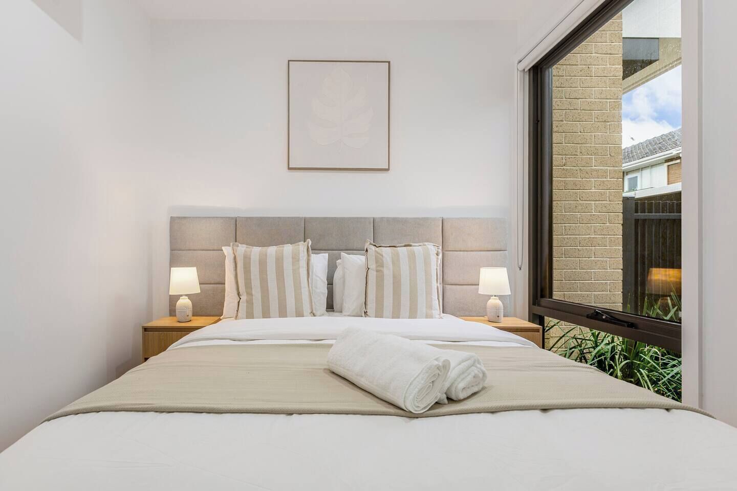 A hotel room with a large bed, two bedside tables with lamps, a framed picture on the wall, and a window with a view of greenery.