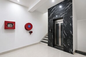 A modern interior with a marble wall, a red fire alarm, and a staircase leading to an elevator. - FabHotel Shiv Shakti Dham (Vrindavan)
