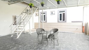 Un espacio al aire libre techado con una escalera, dos sillas de mimbre y plantas en macetas colgantes.