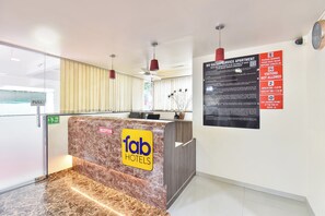 Zona de recepción de un hotel, con mostrador de mármol, una mesa de recepción y un tablón informativo en la pared.