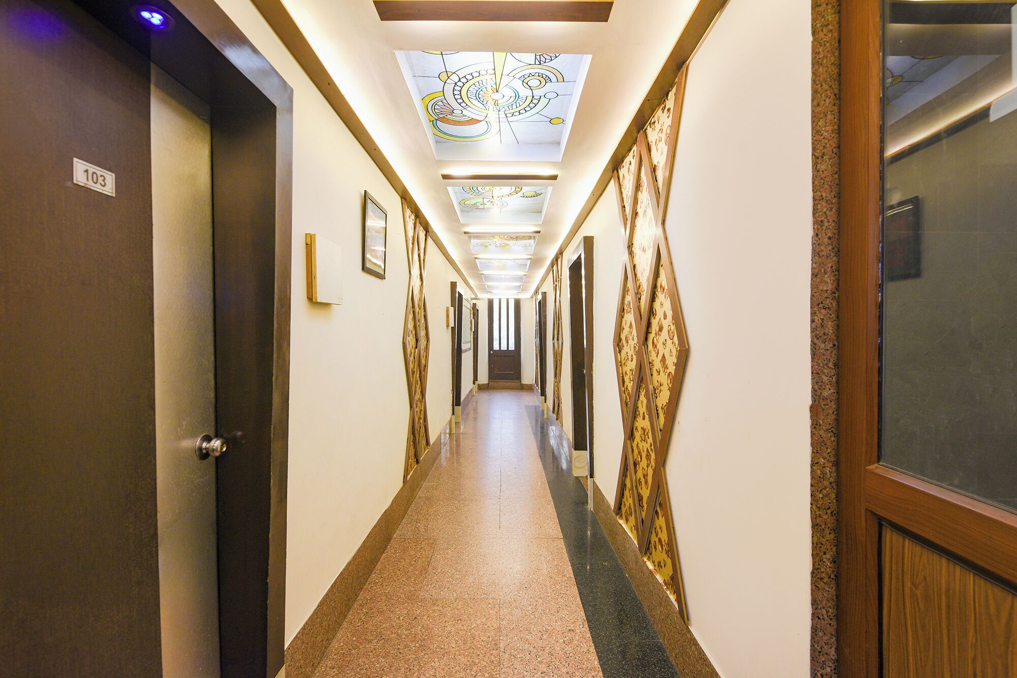 Ein Hotelgang mit Oberlicht, gerahmten Kunstwerken und einer Tür mit der Nummer 103.
