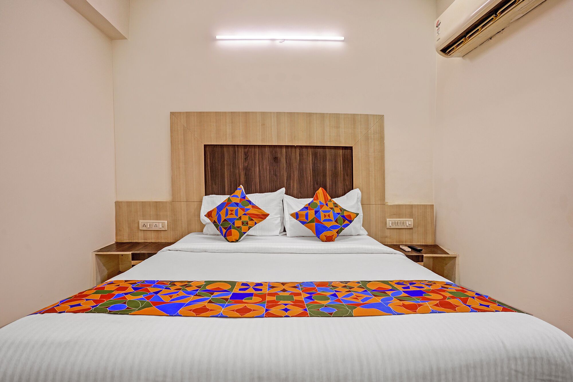 Habitación de hotel con cabecera de madera, cama con ropa blanca, dos almohadas decorativas, mesitas de noche con lámparas y un equipo de aire acondicionado.