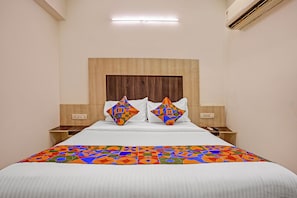A hotel room with a wooden headboard, a bed with white bedding, two decorative pillows, bedside tables with lamps, and an air conditioning unit. - Fabhotel Mid Way (Gandhinagar)