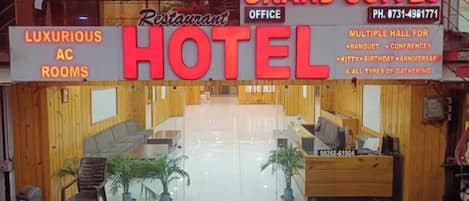 A hotel interior with a reception desk, a waiting area with chairs and a glass partition, and a visible phone number.