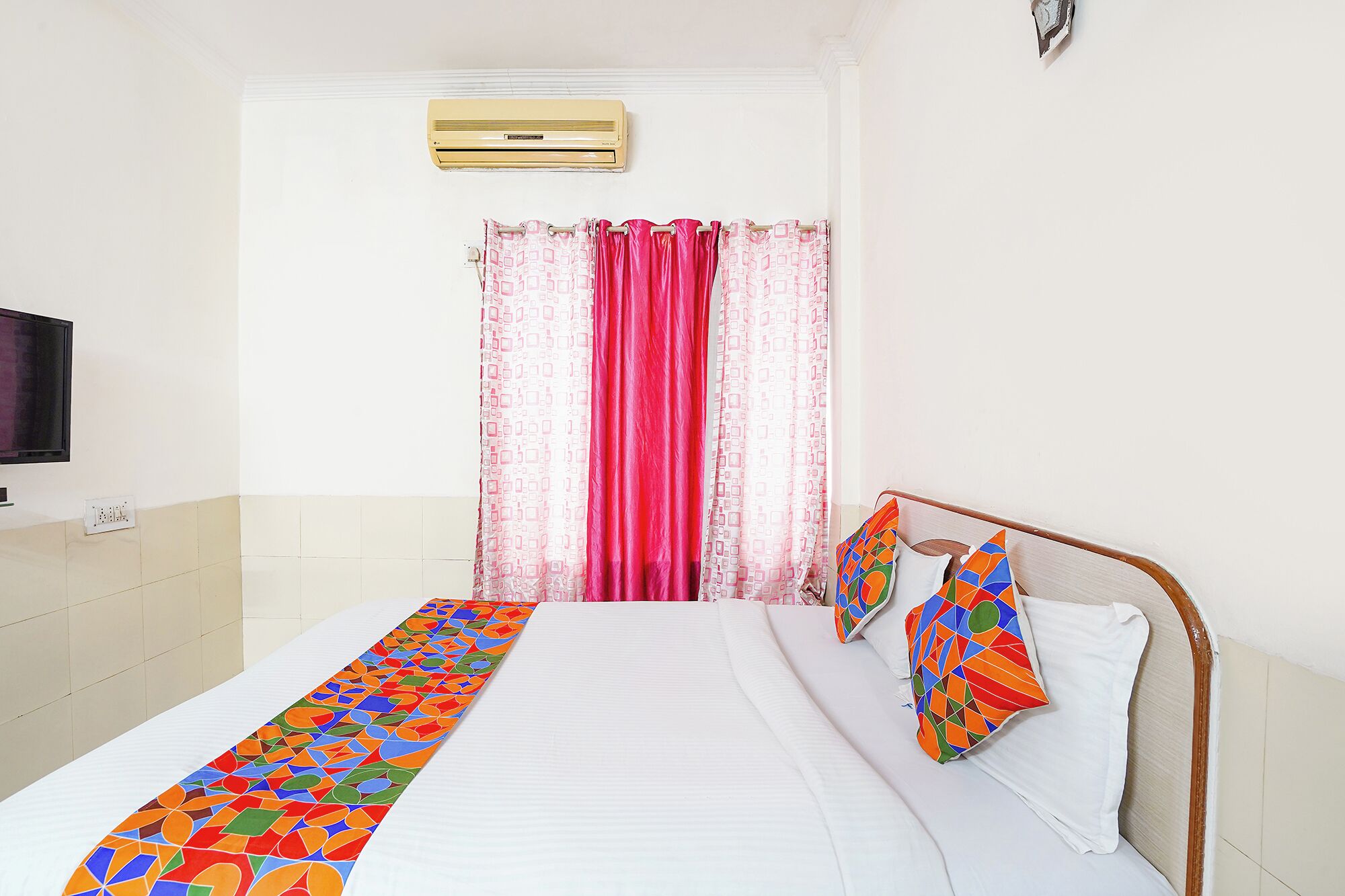 A hotel room with a bed, a TV, a window with curtains, and a wall-mounted air conditioner.