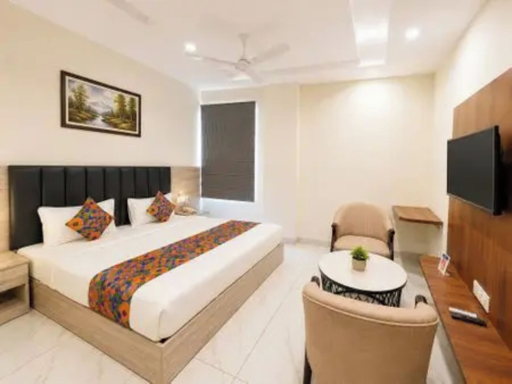 Habitación de hotel con cama, una silla, una mesita, televisión y un cuadro en la pared.