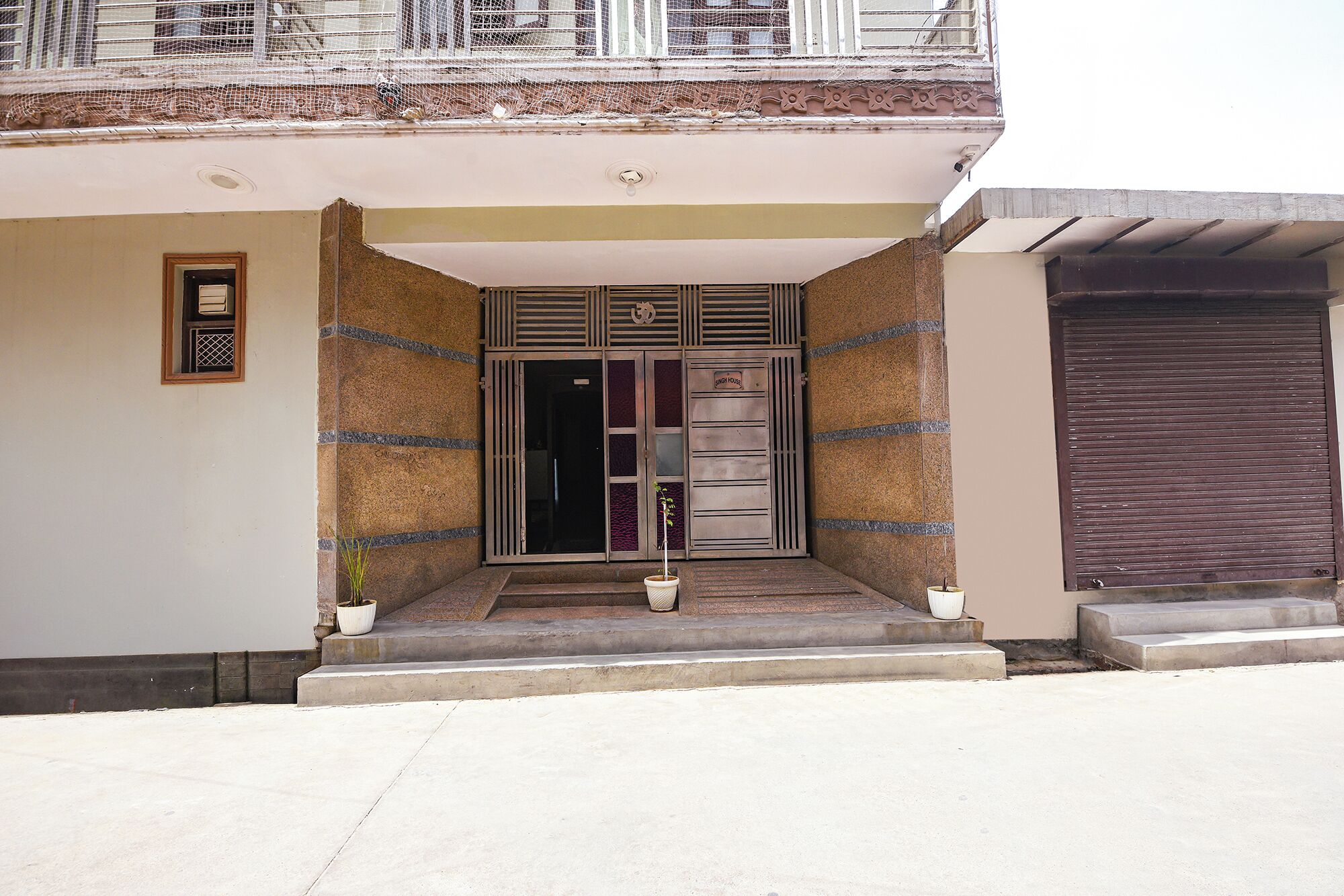 A building entrance with a metal gate, a small potted plant, and a closed shutter to the right.