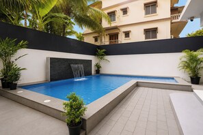 A swimming pool with a waterfall feature, surrounded by potted plants and a building in the background.