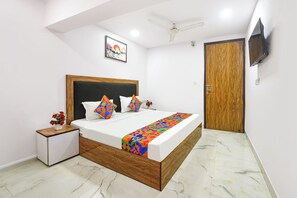 A bedroom with a wooden bed frame, white bedding, a colorful geometric throw, a small bedside table with a vase of flowers, a ceiling fan, and a wooden door. - FabHotel Samrat Residency (New Delhi)