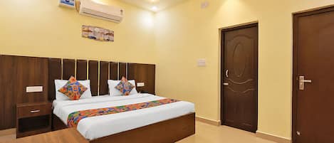 A hotel room with a bed, bedside tables, an air conditioning unit, a door, and a wall-mounted light fixture.