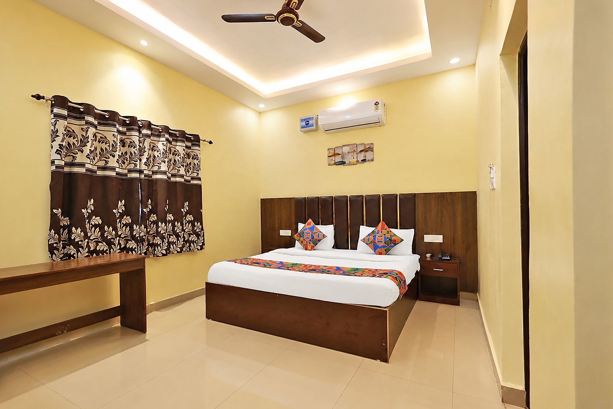 A hotel room with a bed, bedside table, nightstand, air conditioning unit, ceiling fan, and a window with curtains.