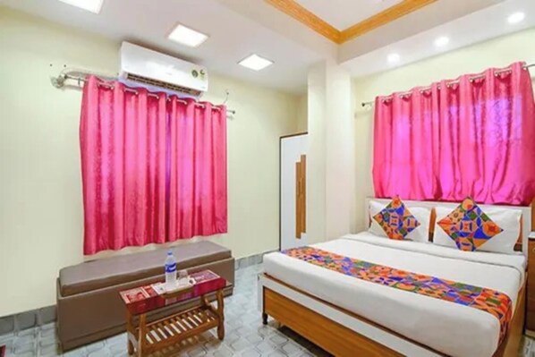 A hotel room with a bed, a bedside table, a small table with a water bottle, and a window with curtains. - Via Om Shanti International (Kolkata)