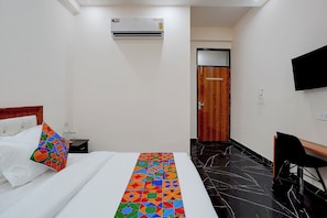 A hotel room with a bed, a colorful pillow, a bedside table with a glass, an air conditioning unit, a wooden door, and a black floor. - FabHotel Luxora (Noida)