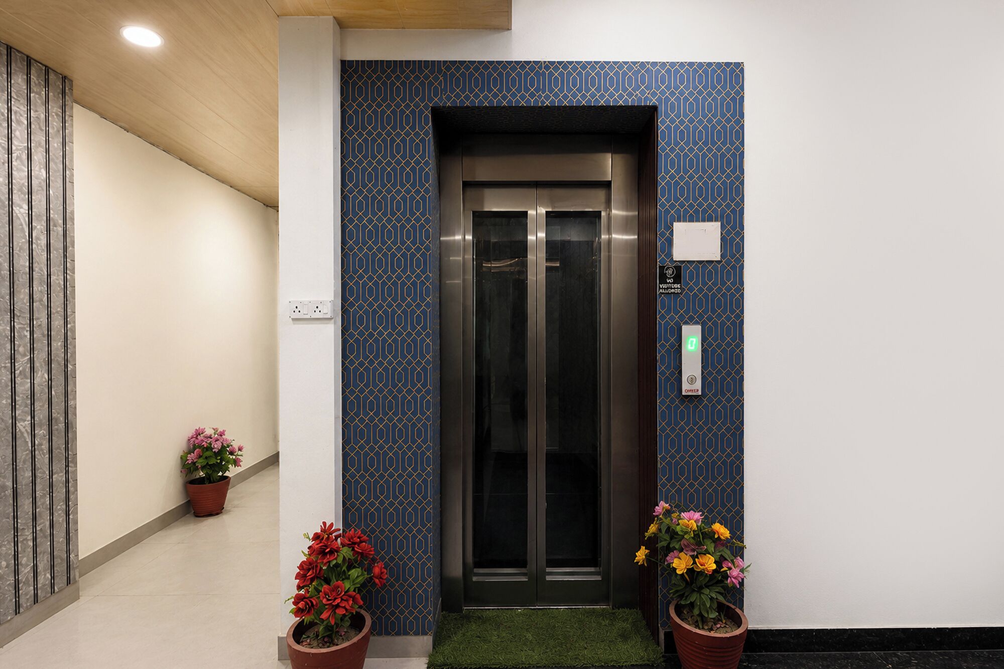 Un ascensor amb una paret de disseny geomètric, un tapet verd a l'entrada i plantes en testos amb flors vermelles i grogues.