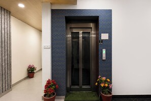 Un ascensor con pared de diseño geométrico, felpudo verde en la entrada y plantas en macetas con flores rojas y amarillas.