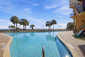 A clear swimming pool with a tiled edge, surrounded by lounge chairs and palm trees, with a building featuring a balcony on the right. - 1 BR Oceanfront Pet Friendly Fall Upgrades! (Myrtle Beach)