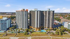 Un hotel alt amb múltiples balcons, una piscina i una zona enjardinada amb palmeres.