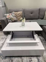 Uma mesa de centro branca com um vaso de flores, um sofá cinzento e uma almofada estampada.