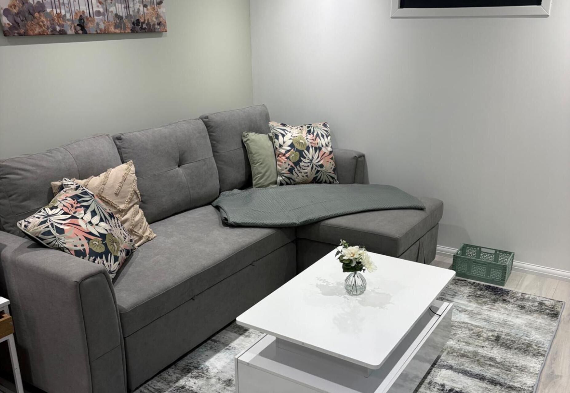 Un sofá seccional gris con cojines florales, una mesa de centro blanca con un florero y una alfombra gris.