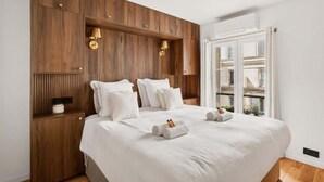 A bedroom with a wooden headboard, white bedding, and a window with a view of buildings. - In the heart of Paris, new flat for 6, with AC (Paris)