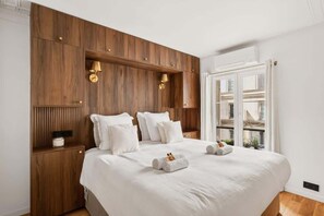 Ein Schlafzimmer mit einem hölzernen Kopfende, weißer Bettwäsche und einem Fenster mit Blick auf Gebäude.