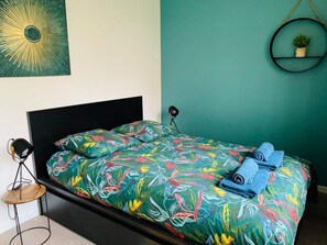 Ein Bett mit bunter Blumenbettdecke, einem schwarzen Kopfteil und einem kleinen Topfpflanzen auf einer türkisblauen Wand.