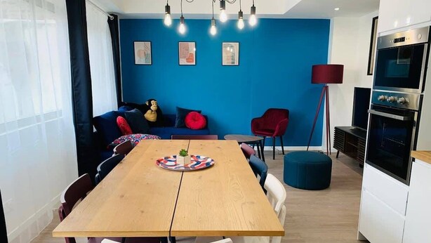 Une cuisine moderne dotée d’un four encastré, un coin repas avec une table en bois et des chaises blanches, et un mur d’accent bleu.