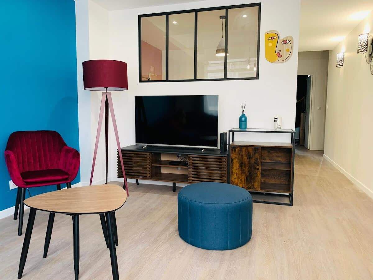 A modern living room with a red chair, a wooden coffee table, a blue ottoman, a TV on a wooden stand, and a small cabinet with a vase.