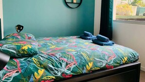 Un lit recouvert d’une couvre-lit aux motifs floraux colorés, une petite étagère avec une plante et une fenêtre donnant sur un paysage verdoyant.