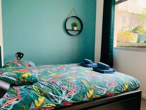 A bed with a colorful floral bedspread, a small shelf with a plant, and a window with a view of greenery. - Beautiful 3-bedroom apartment / Porte de Versailles (Paris)
