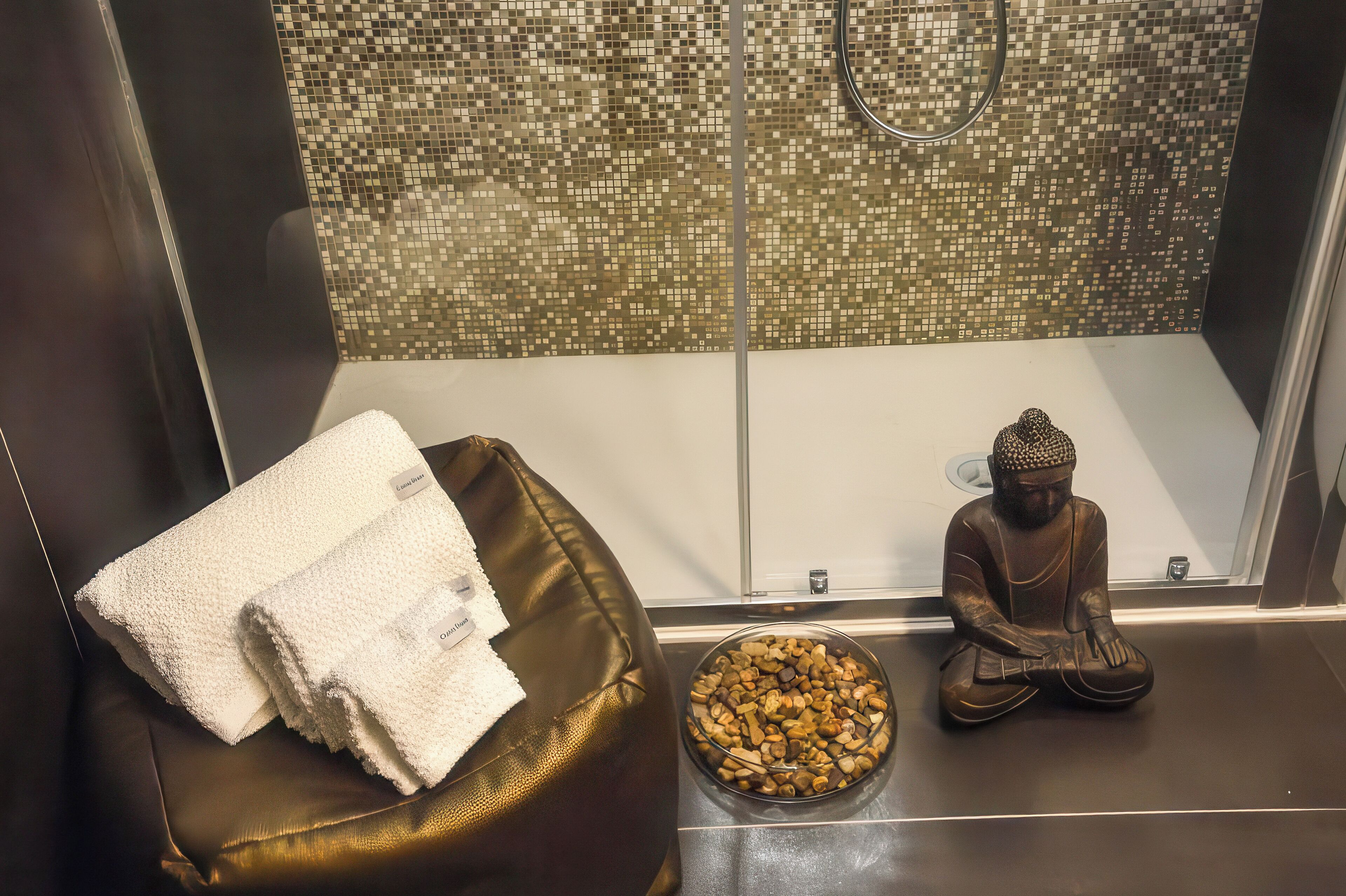 A bathroom with a mosaic-tiled shower wall, a brown leather bench with white towels, and a Buddha statue.