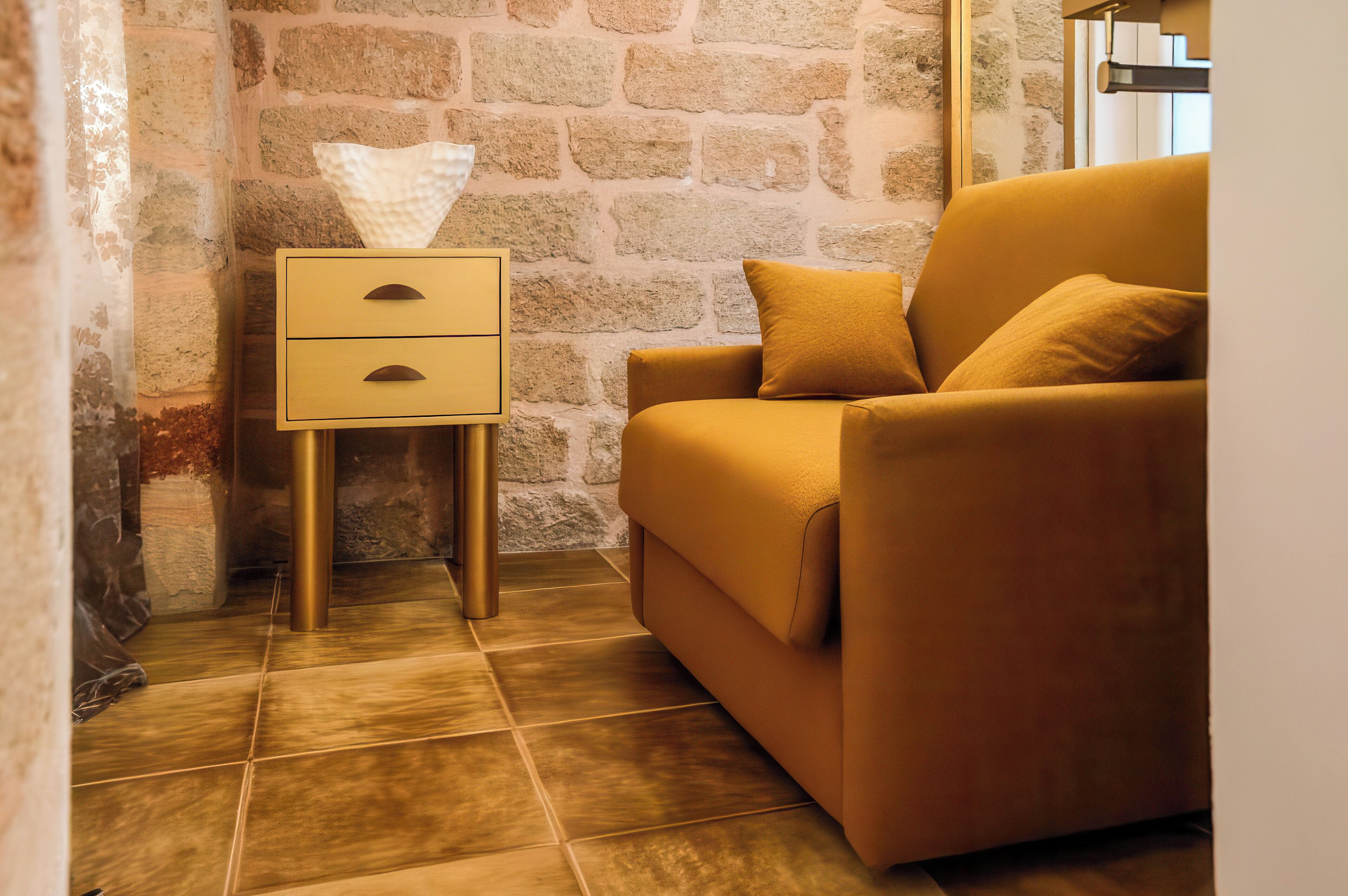 A room with a stone wall, a yellow sofa, a bedside table with a tissue box, and a lamp.