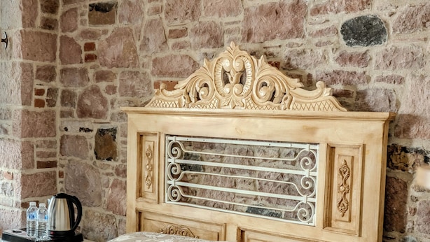 Un dormitorio con pared de piedra, cabecera de madera, cama con ropa de cama estampada, mesita de noche con un control remoto y una cafetera en la mesita auxiliar.