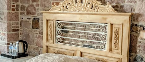 Un dormitorio con pared de piedra, cabecera de madera, cama con ropa de cama estampada, mesita de noche con un control remoto y una cafetera en la mesita auxiliar.