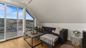 Ein modernes Schlafzimmer im Loftsstil mit einem großen Oberlicht, einem dunkelgrauen Sofa, einem kleinen runden Tisch und einer gemütlichen Sitzecke.