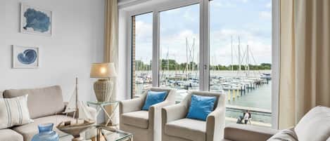 Una sala de estar moderna con vista a un puerto deportivo, que incluye un conjunto de sofás, una mesa de centro de vidrio y elementos decorativos.