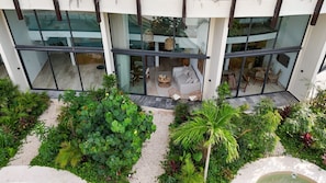 Ein modernes Gebäude mit großen Glasfenstern, einer Sitzecke mit Sofa und Sesseln und einem gepflegten Garten mit verschiedenen Pflanzen und einem kleinen Wasserspiel.