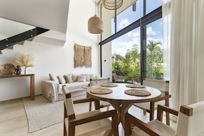 Ein modernes Wohnzimmer mit einem runden Holztisch, einem weißen Sofa und großen Fenstern mit Blick auf Grünflächen.