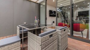 Uma área externa moderna com mobiliário de vime, uma mesa de vidro e uma televisão fixada na parede.