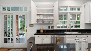 A kitchen with white cabinets, a black countertop, and a window with a view of outdoor furniture. - The Sanctuary at Morris Creek | 4BR Luxury Stay (Charles City)