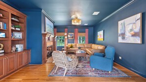 A room with a blue wall, a wooden floor, a brown sofa, a blue armchair, a wicker chair, a coffee table, and a bookshelf. - The Sanctuary at Morris Creek | 4BR Luxury Stay (Charles City)