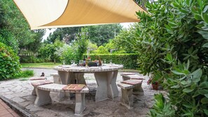 Una mesa redonda de piedra con bancos, rodeada de follaje y un sendero de piedra.