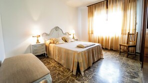 Un dormitorio con cama, mesitas de noche, una silla y una ventana con cortinas.
