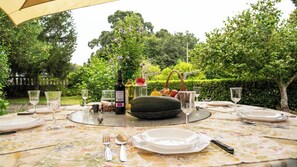 Mesa de comedor al aire libre dispuesta con platos, cubiertos, copas de vino y botellas, rodeada de abundante vegetación.