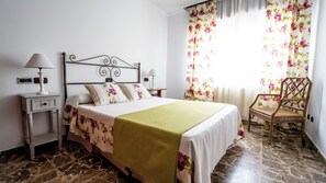 Ein Schlafzimmer mit einem Bett, zwei Nachttischen, einem Sessel und einem Fenster mit blumigen Vorhängen.