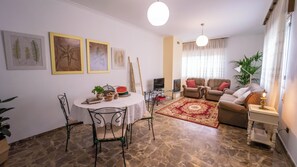 Ein Wohnzimmer mit einer Sofagarnitur, einem Esstisch mit Stühlen, einem Fernseher und Bildern an der Wand.