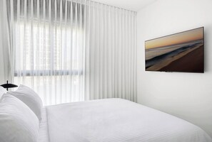 Quarto de hotel com uma cama, uma janela com cortinas leves e uma TV na parede exibindo uma paisagem de praia.