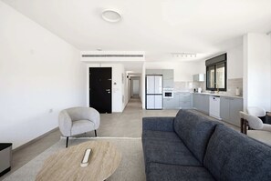 Ein modernes Wohnzimmer mit einem grauen Sofa, einem runden Couchtisch und einem grauen Sessel. Im Hintergrund ist eine Küche mit weißen Geräten und Schränken zu sehen.